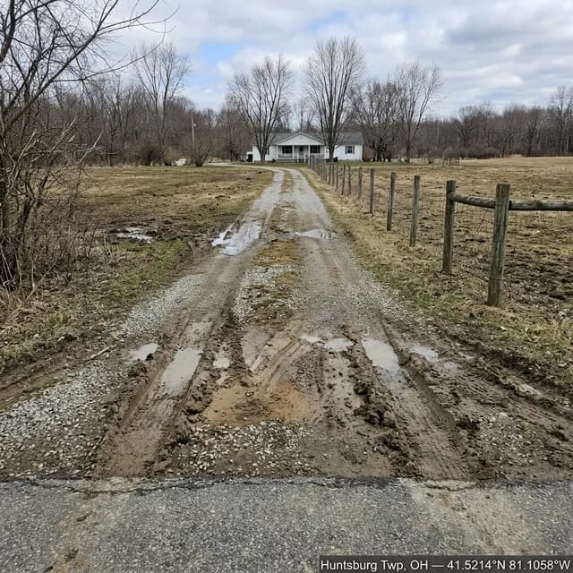 Muddy gravel driveway in Huntsburg Township, Ohio before restoration — rutted wheel tracks and water collecting along both edges beside a split-rail fence