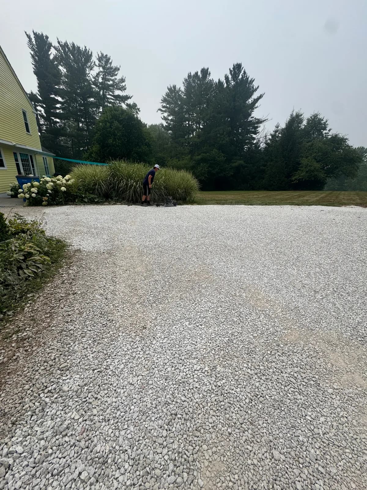 Finished gravel parking pad beside a yellow house, with a crew member mowing along the edge