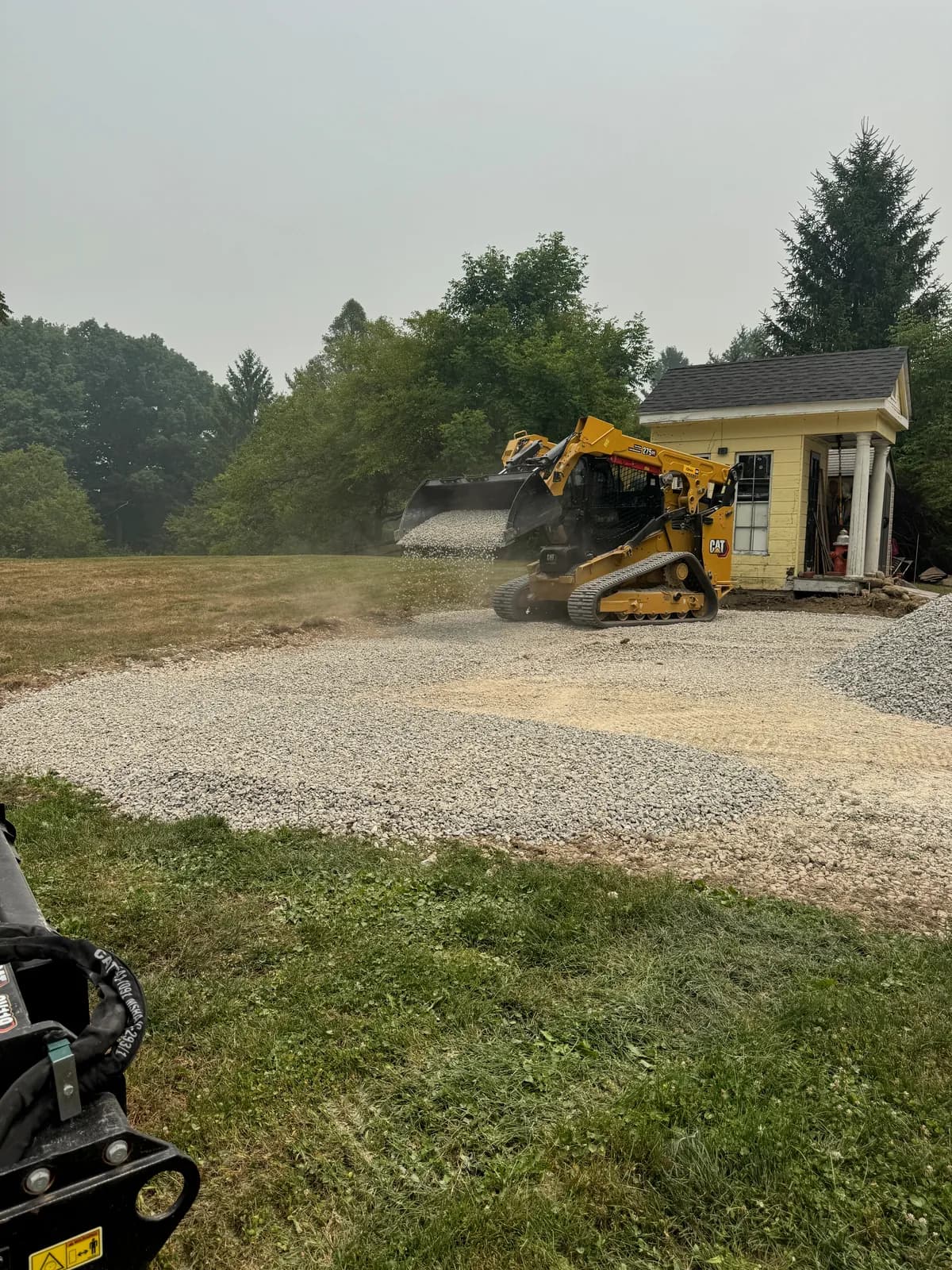 Track loader spreading new gravel across a parking pad beside a small yellow building, with a pile of stone to one side