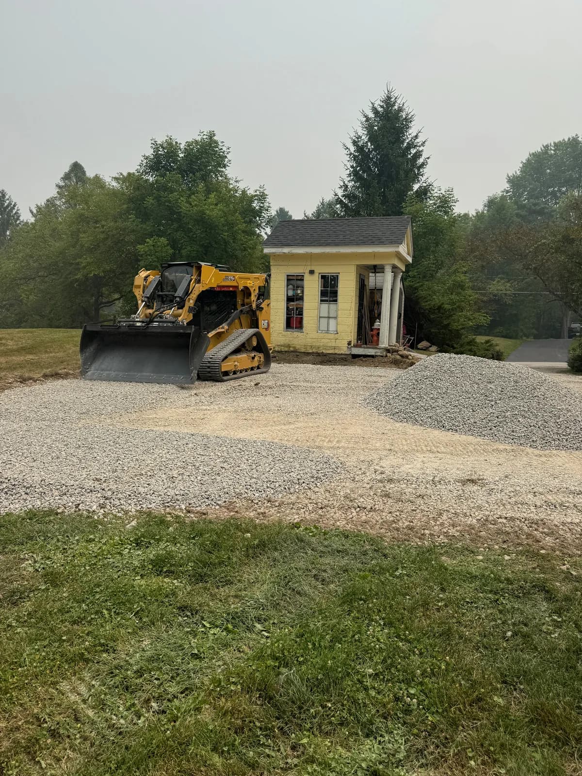 Track loader beside a small yellow building and a pile of new gravel, on a partly spread parking pad