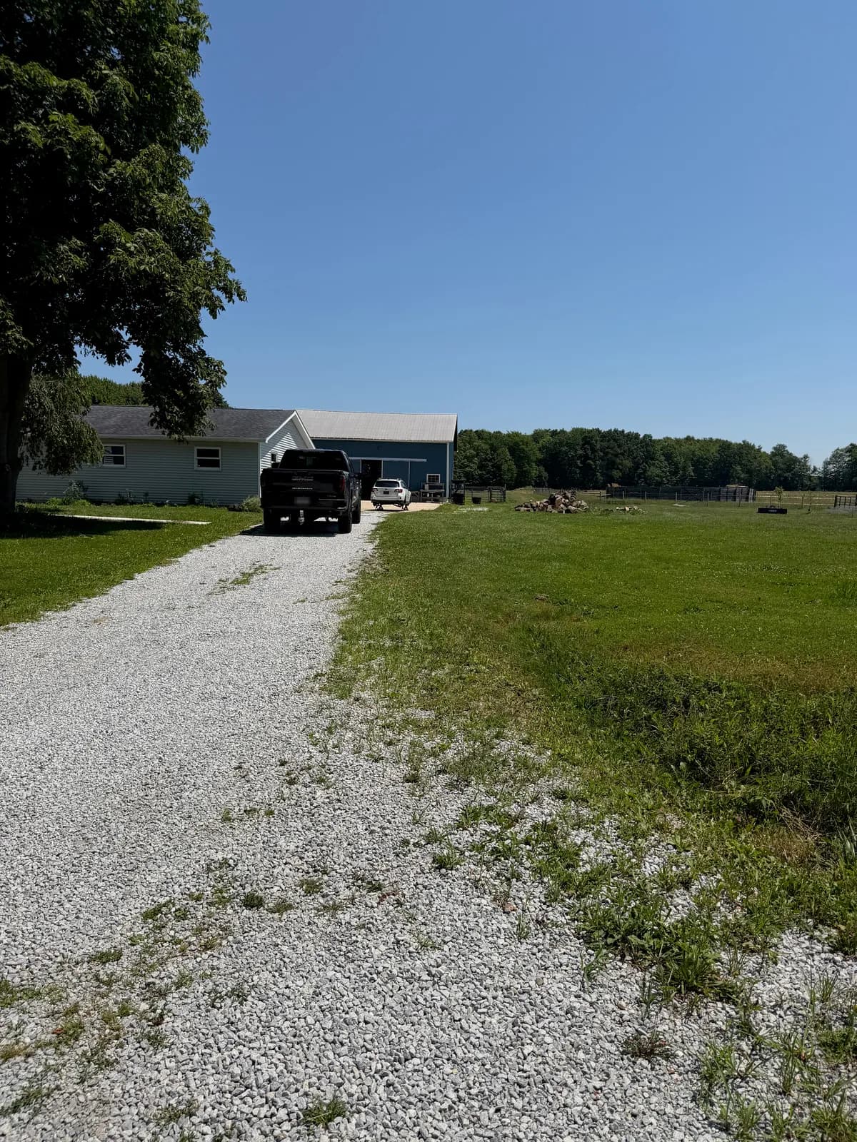 Gravel driveway leading to a light-blue house and barn before GravelBoss work, with grass and weeds growing through the stone along its edges